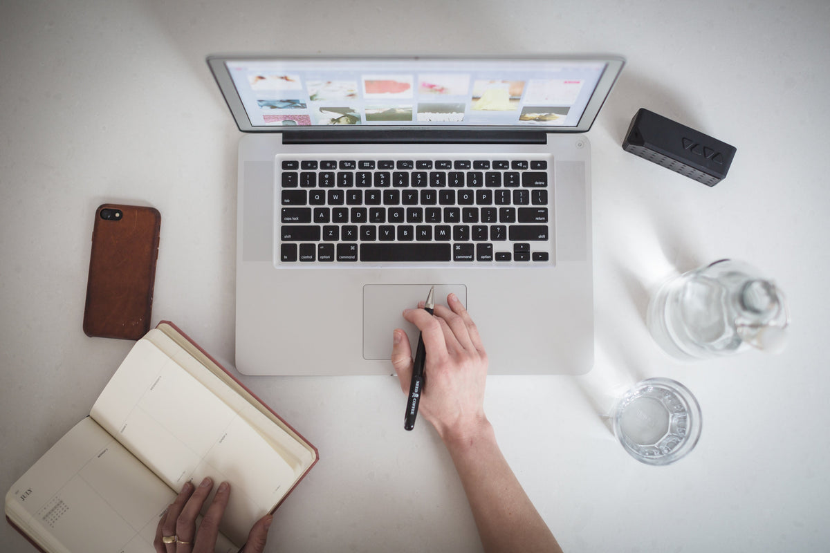 Overhead view of a person using a laptop beside an open notebook, phone and glasses of water on a white desk.